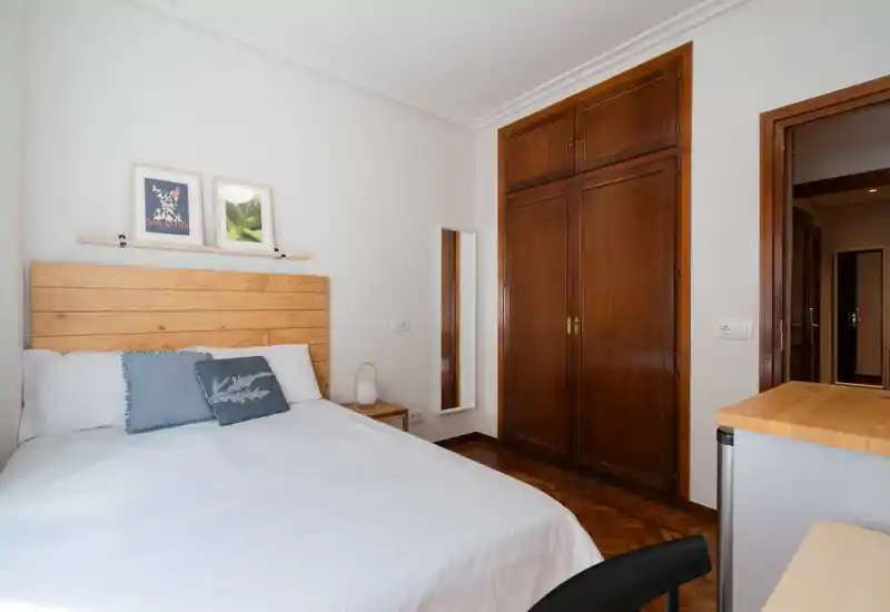 Dormitorio luminoso y ordenado con cabecero de madera, ropa de cama blanca y cojines decorativos. Armario empotrado de madera y una lámpara de mesita aportan calidez al espacio.