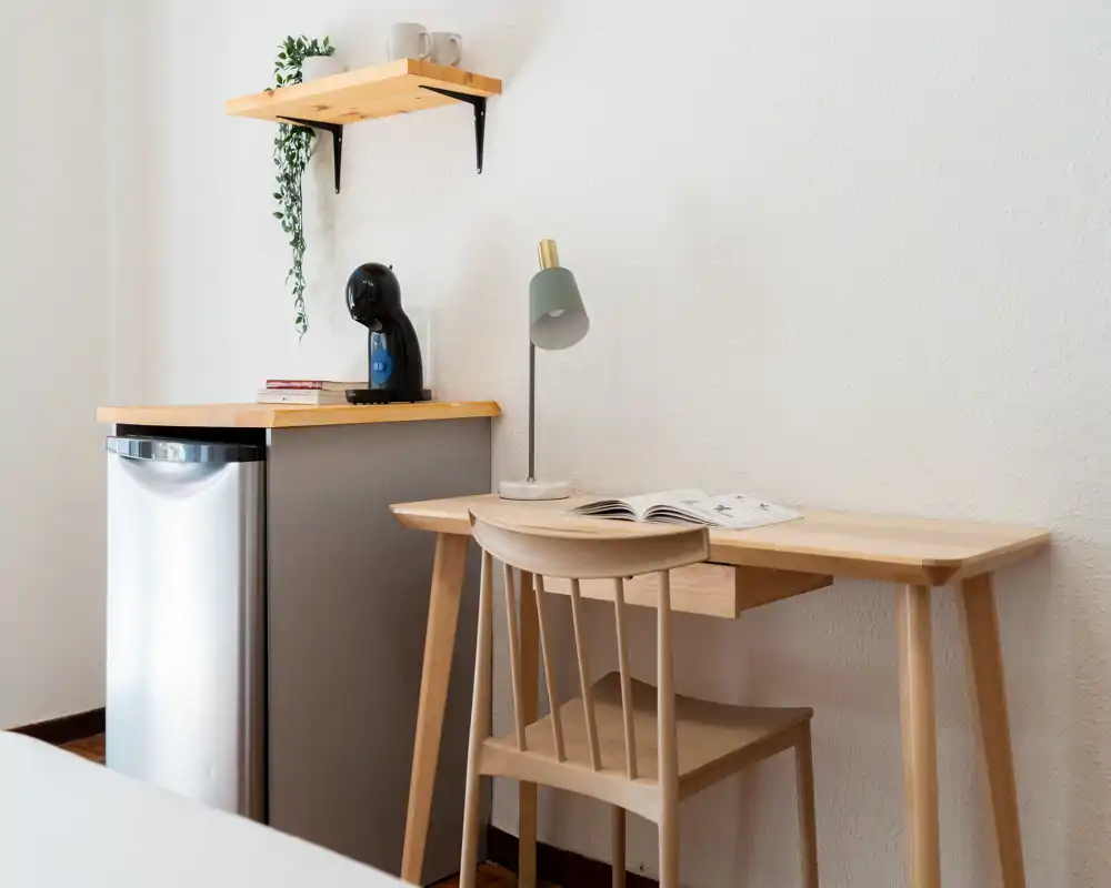 Rincón de dormitorio compacto con escritorio de madera, silla, mini nevera y lámpara; paredes neutras y decoración minimalista crean un espacio de trabajo tranquilo en la habitación.