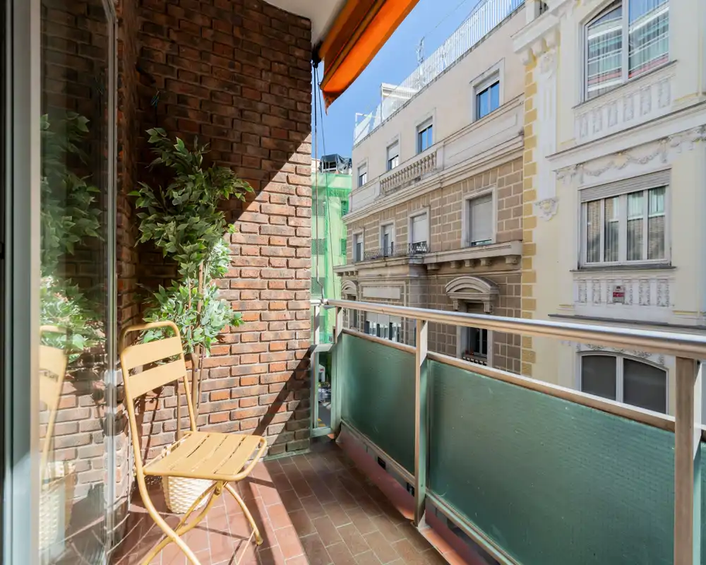 Balcón urbano soleado con una pequeña silla, pared de ladrillo y barandilla de vidrio — ideal para el café de la mañana con vistas a la calle.