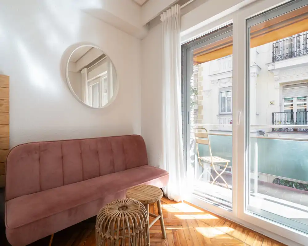Zona de estar luminosa y acogedora con sofá de terciopelo rosa, luz natural de las puertas correderas y un pequeño balcón — ambiente aireado y relajante.