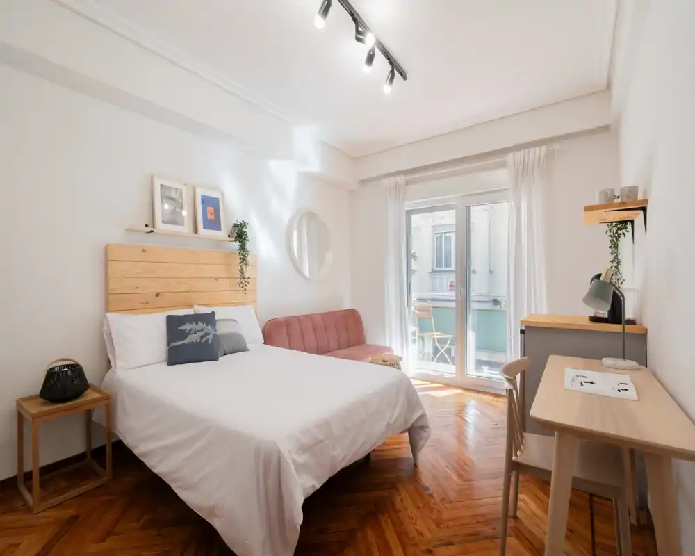 Dormitorio luminoso y acogedor con cabecero de madera, ropa de cama blanca y una pequeña zona de estar junto al balcón. La luz natural entra por las puertas de cristal; hay escritorio y silla para trabajar.