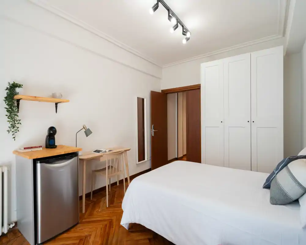 Dormitorio luminoso y ordenado con cama doble, armario blanco y pequeño escritorio. Suelo de parquet cálido, ropa de cama acolchada y un mini-frigorífico hacen la estancia cómoda.