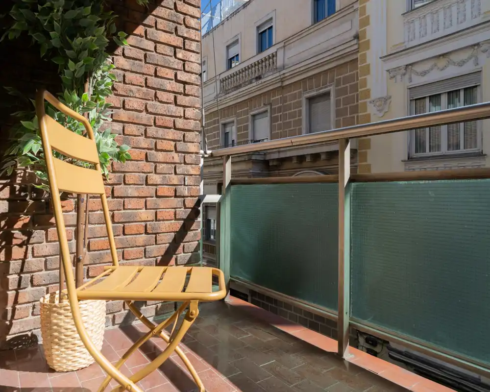 Pequeño balcón soleado con una silla plegable amarilla, pared de ladrillo, planta en maceta y vidrio traslúcido en la barandilla — ideal para tomar un café por la mañana en la ciudad.
