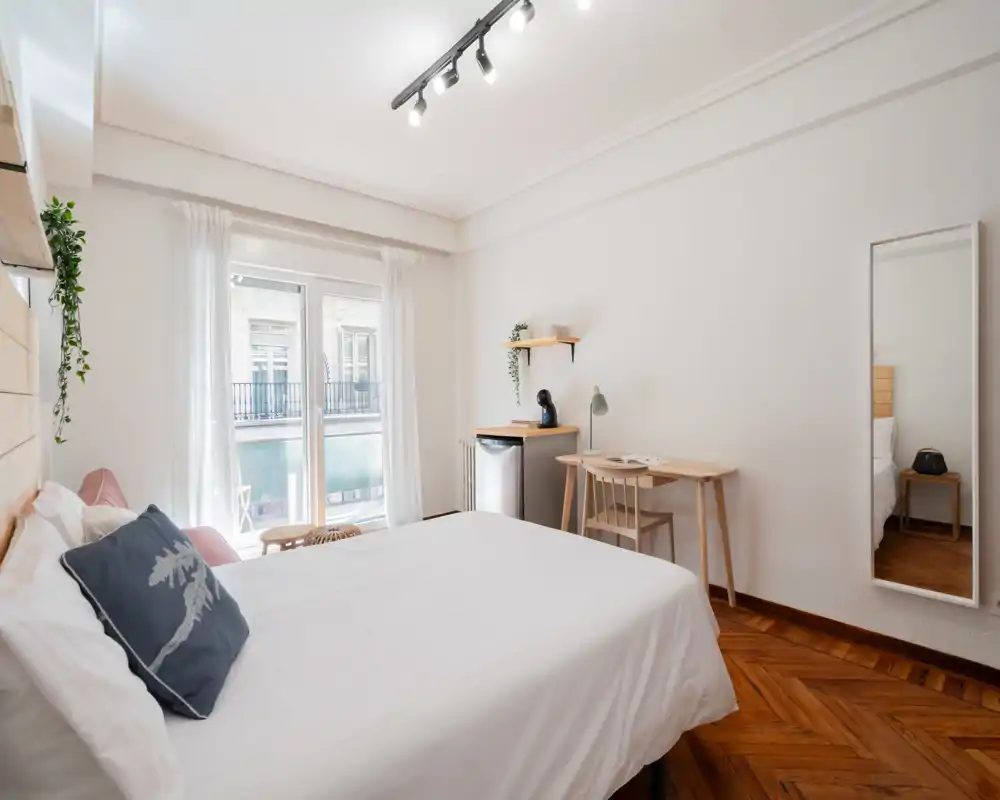 Dormitorio moderno y luminoso con cama grande, suelo de parquet y escritorio junto a la ventana. Textiles suaves, plantas e iluminación equilibrada crean un ambiente acogedor, ideal para estancias cortas o largas.