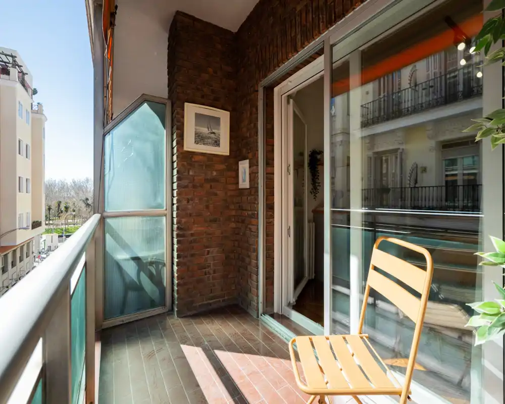 Balcón soleado amueblado con una silla metálica, puerta corredera de cristal al interior y vista a la calle y edificios vecinos — perfecto para el café de la mañana.