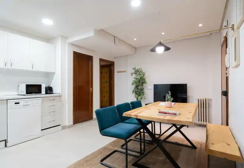 Zona de comedor moderna y luminosa con mesa de madera, banco y sillas turquesa; abierta a una cocina compacta y decorada con una planta y una lámpara colgante para un ambiente acogedor.
