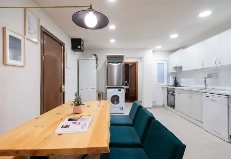 Cocina moderna y luminosa con una larga mesa de madera y sillas tapizadas color teal; electrodomésticos integrados (horno, lavavajillas, nevera) y lavadora visible, espacio práctico y acogedor.