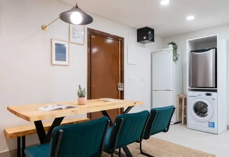 Área de comedor moderna y luminosa con mesa de madera maciza y banco, sillas tapizadas en verde azulado y electrodomésticos integrados a un lado — acogedora y bien decorada para un piso compacto.