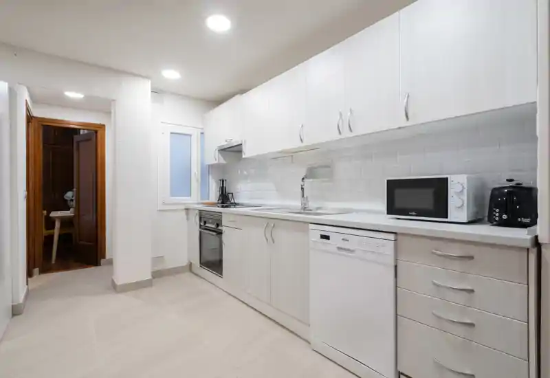 Cocina moderna en galería, luminosa, con muebles claros, horno empotrado, lavavajillas y pequeños electrodomésticos; salpicadero tipo metro y suelo neutro.
