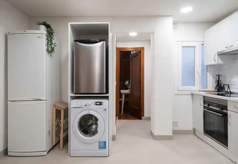 Cocina moderna y compacta con electrodomésticos visibles: frigorífico, lavadora y horno, suelo de azulejos claros y armarios blancos que ofrecen un espacio de cocina limpio y funcional.