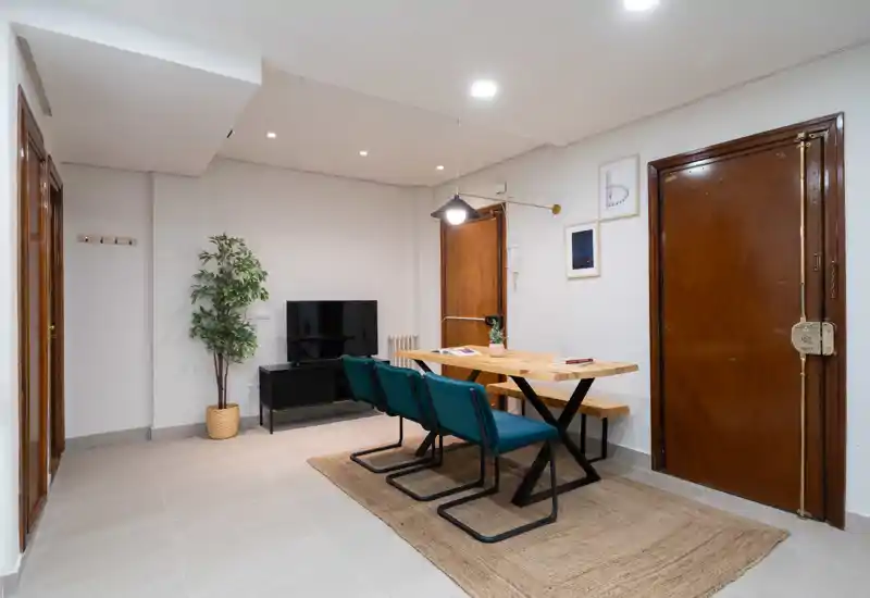 Zona de estar/comedor compacta y moderna con mesa de madera, banco y sillas turquesa sobre una alfombra, mueble TV y planta decorativa — espacio luminoso y bien presentado, ideal para estancias cortas.