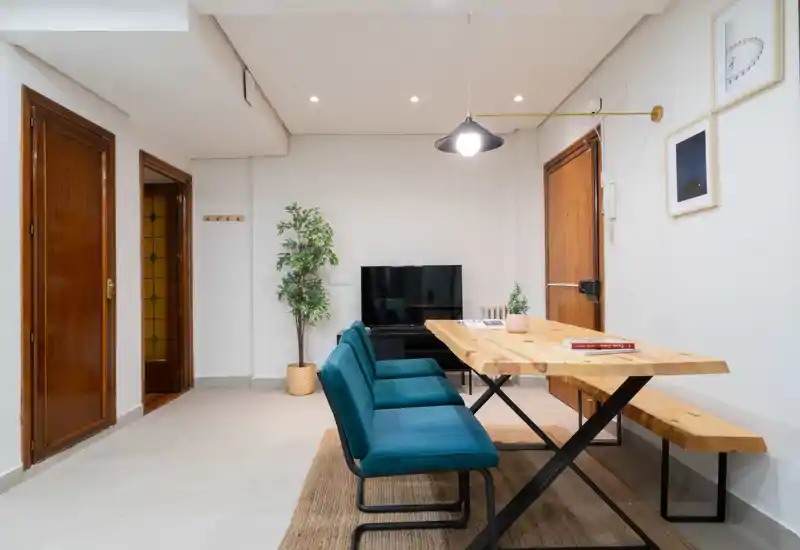 Zona de día luminosa y moderna con mesa de madera, banco y sillas tapizadas en turquesa, mueble TV y planta decorativa: perfecta para comidas informales y reuniones pequeñas.