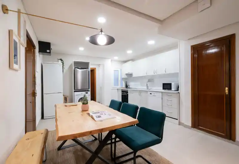 Cocina moderna de concepto abierto, luminosa, con mesa de madera natural, banco y sillas tapizadas; armarios blancos y electrodomésticos que crean un ambiente acogedor para cocinar y comer.