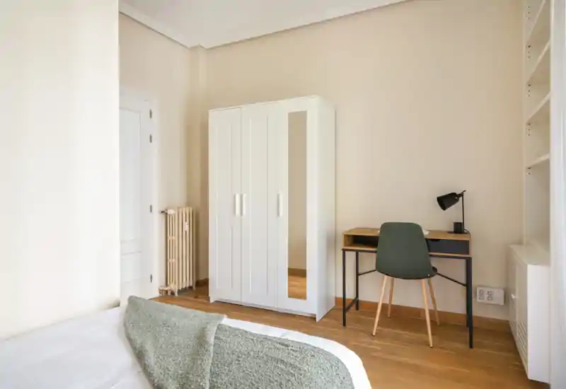Bright, minimal bedroom with a white wardrobe, bedside bed partially visible in the foreground and a small desk with lamp — warm wooden floors and neutral walls create a calm atmosphere.