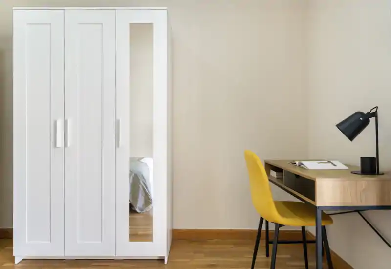 A minimal bedroom corner featuring a white wardrobe with a full-length mirror reflecting the edge of a bed, and a small wooden desk with a yellow chair and task lamp. Clean, modern styling and soft natural light create a calm workspace within the sleeping area.