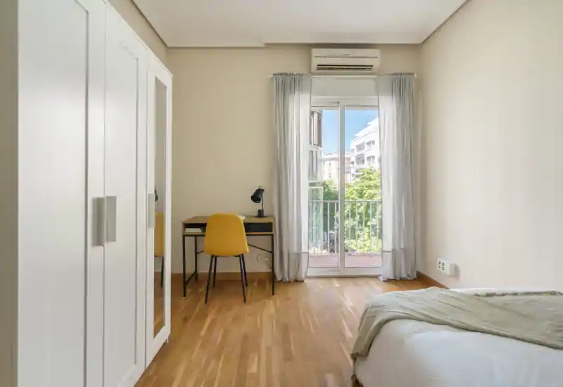 Bright, minimally decorated bedroom with wooden floors, a visible double bed with light linens, a wardrobe and a small desk facing a balcony with natural light.