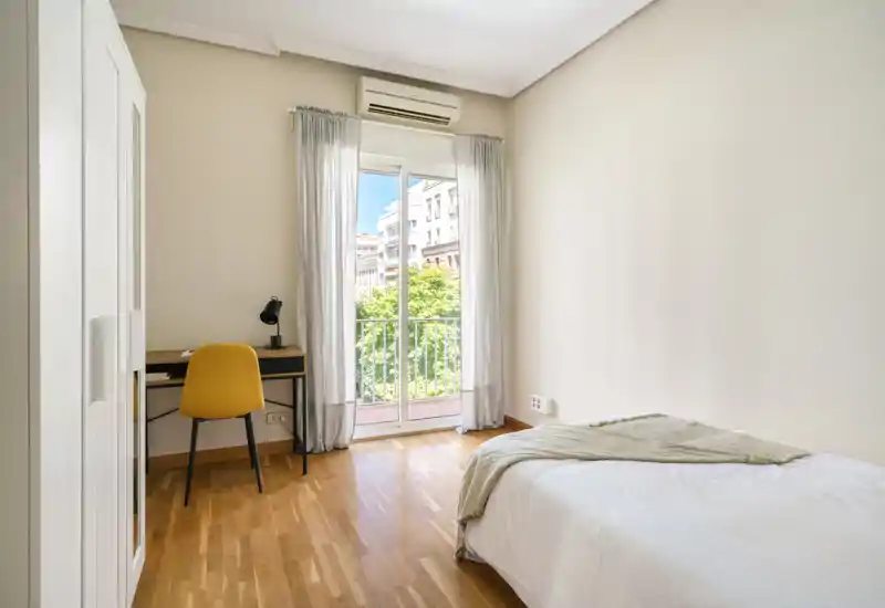 Bright, minimalist bedroom with a single bed, wooden floor and a desk beside a large balcony door that fills the room with natural light.