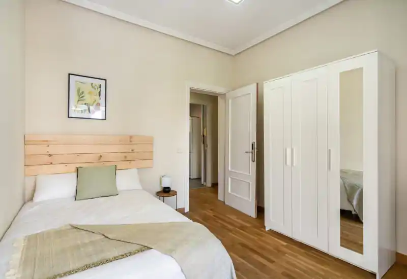 Bright, minimalist bedroom with a wooden headboard, neatly made double bed, bedside table and a white wardrobe with mirror—ideal for a clean, modern rental listing.