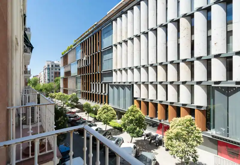Vista desde el balcón a una calle urbana con árboles y la fachada de un edificio moderno, tomada a plena luz del día. Perfecta para mostrar la orientación del piso y la arquitectura cercana.