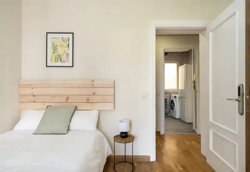 Bright, minimalist bedroom with a wooden slat headboard, white bedding and a small round bedside table with a lamp; neutral tones and simple decor create a calm, welcoming atmosphere.