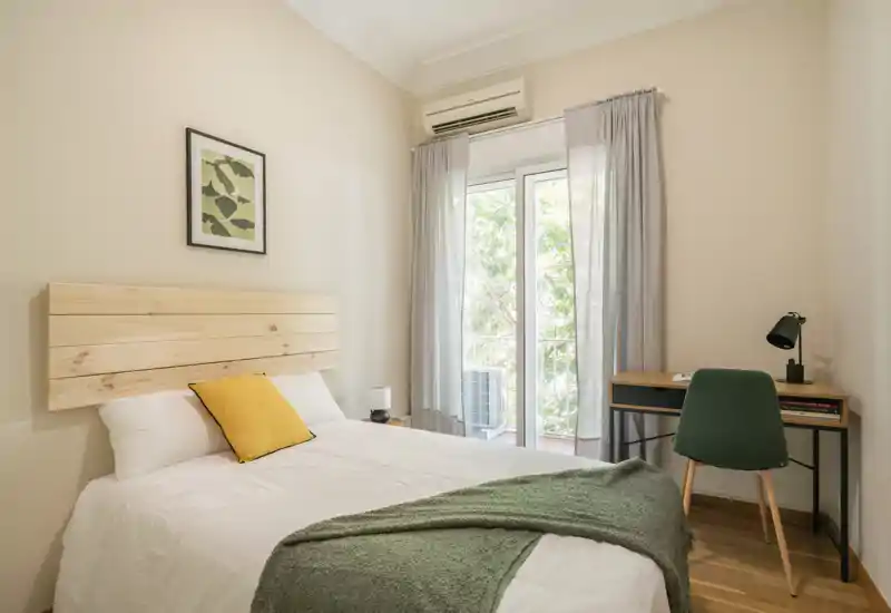 Camera da letto luminosa e minimal con letto matrimoniale, testiera in legno, plaid verde e una piccola scrivania accanto alla porta del balcone che lascia entrare luce naturale.