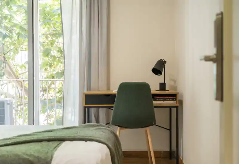 Angolo camera luminoso e minimal: letto visibile in primo piano, una piccola scrivania in legno e una sedia vicino a una grande finestra che diffonde luce naturale.