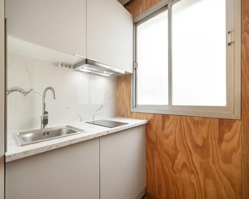 Compact modern kitchen with a stainless sink, induction hob and marble-effect backsplash; warm wood paneling and a large frosted window provide natural light and a cozy atmosphere.