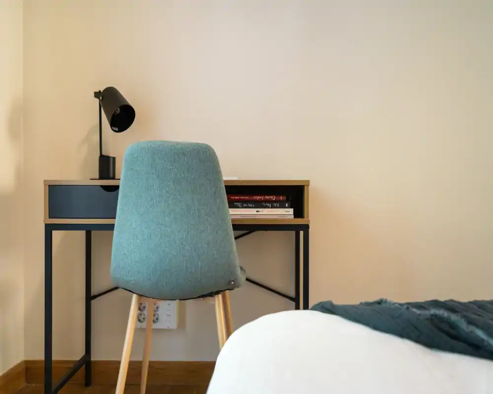 Small, tidy bedroom corner with a desk and lamp beside a partially visible bed. The scene is sharp, simply styled with a teal chair and neutral walls, ideal for a compact apartment listing.
