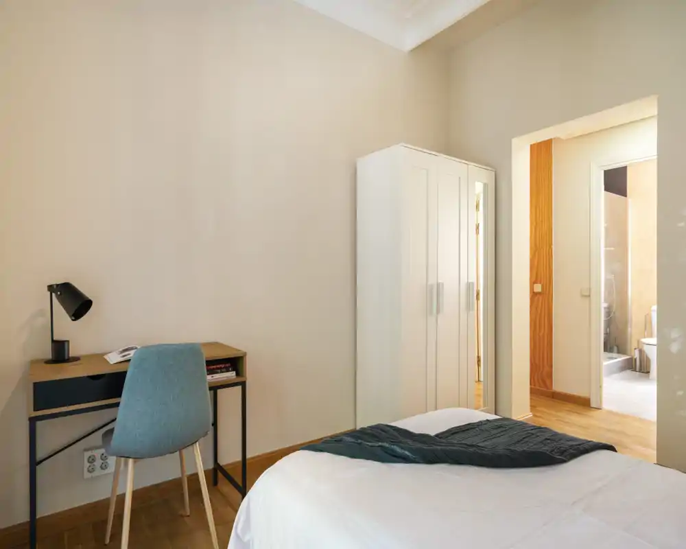 Bright, simple bedroom with a neatly made bed, a small desk and chair, and a white wardrobe. Natural light and warm wood floors give a clean, inviting atmosphere ideal for rental listings.