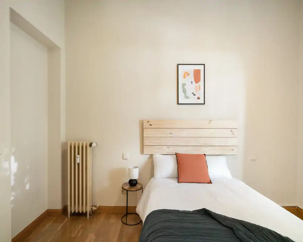 Bright, minimal bedroom with a wooden headboard, white linens and a warm accent cushion; simple bedside table and lamp create a calm, modern feel.