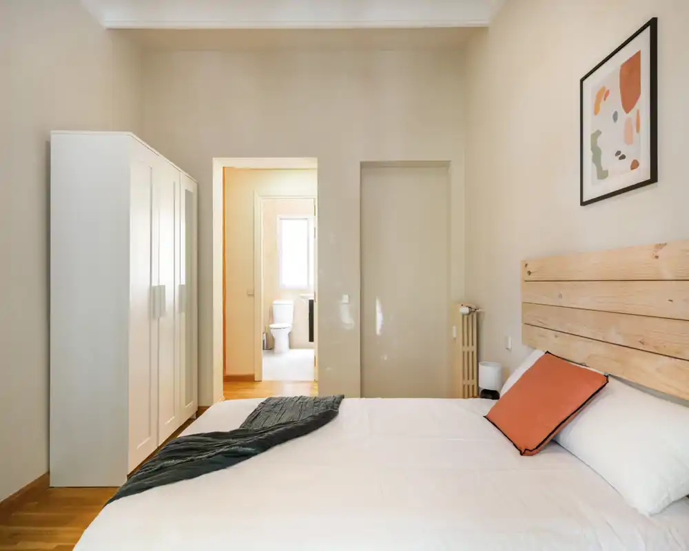 Bright, modern bedroom with a wooden headboard, white wardrobe and a neatly made double bed accented by a coral cushion. Clean lines and warm wood flooring give a calm, welcoming atmosphere.