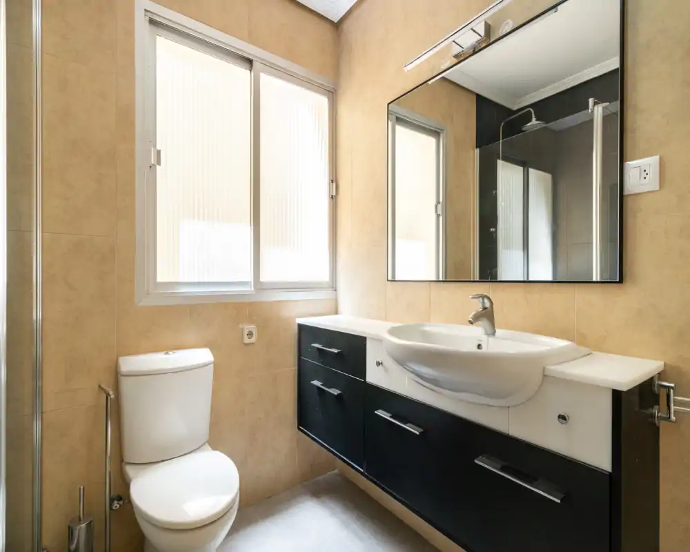 Bright, modern bathroom with a wall-mounted sink and vanity, large mirror and a toilet next to a frosted window providing natural light.