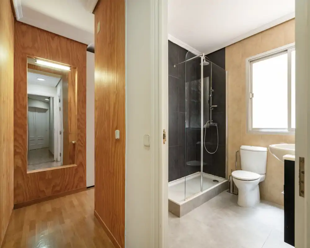 Modern, compact bathroom with a glass-enclosed shower, toilet and sink beside a bright frosted window; neutral tile finishes and clean design.
