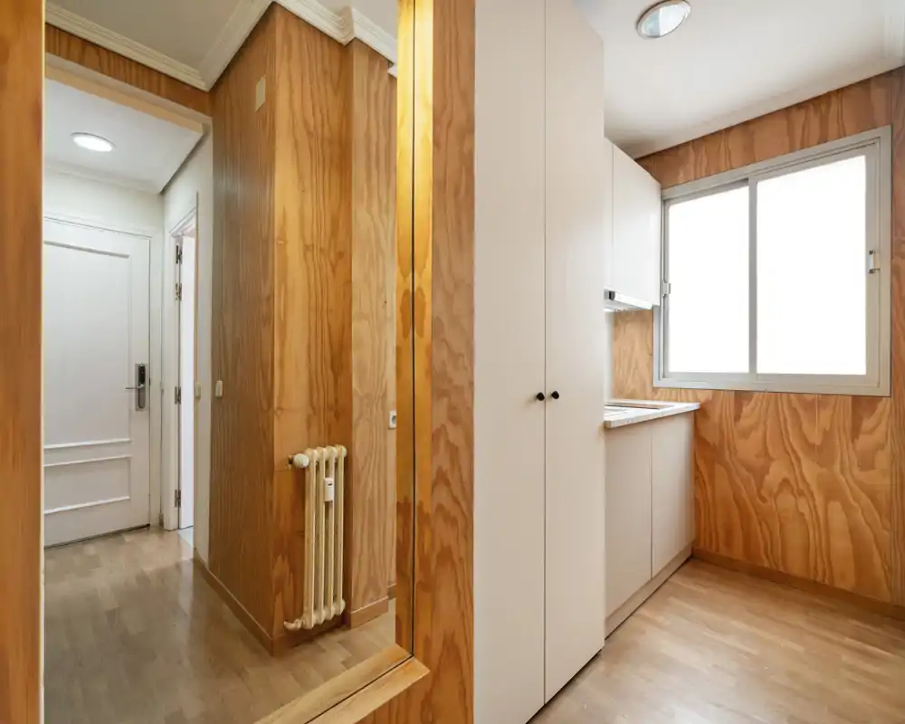 Compact kitchen area with white cabinets, a sink beneath a large window and warm wood-paneled walls that create a cozy, bright nook.