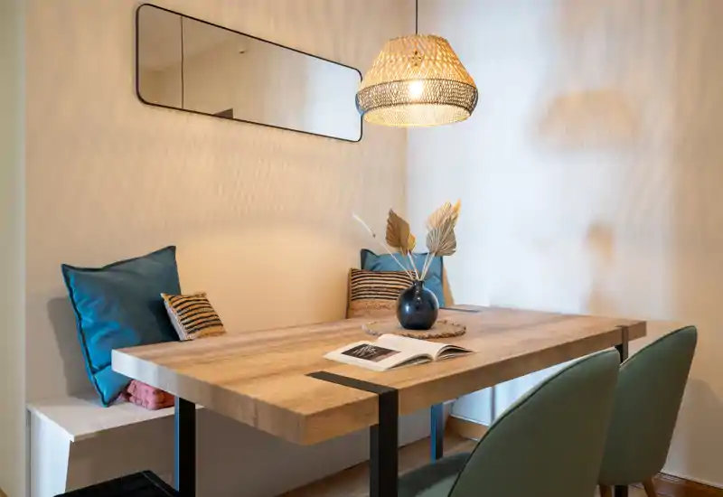 Acogedor rincón de comedor con mesa de madera, cojines en el banco, dos sillas tapizadas y una lámpara de mimbre que genera una atmósfera cálida.