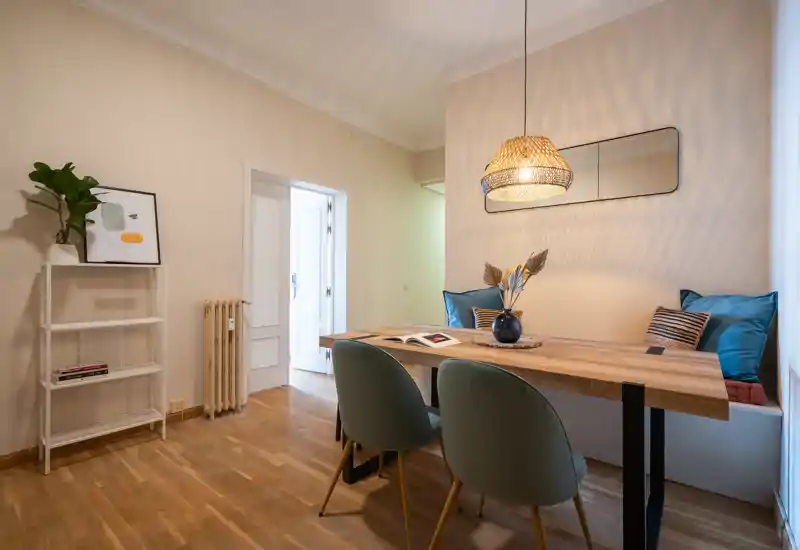 Acogedor comedor con mesa de madera, dos sillas tapizadas y un banco empotrado con cojines, destacado por una lámpara colgante de mimbre y un espejo en la pared.