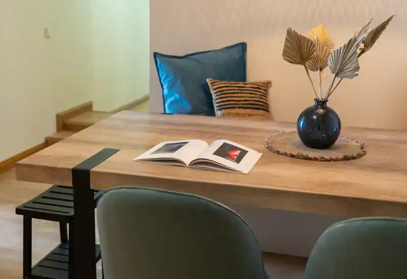 Zona de comedor estilizada con mesa de madera, cojines decorativos y un jarrón con follaje seco — ambiente cálido y bien iluminado, perfecto para un comedor pequeño o rincón de trabajo.