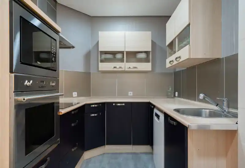 Cocina moderna y luminosa con horno y microondas integrados, fregadero de acero inoxidable y armarios elegantes. Distribución en L compacta, ideal para cocinar con eficiencia.
