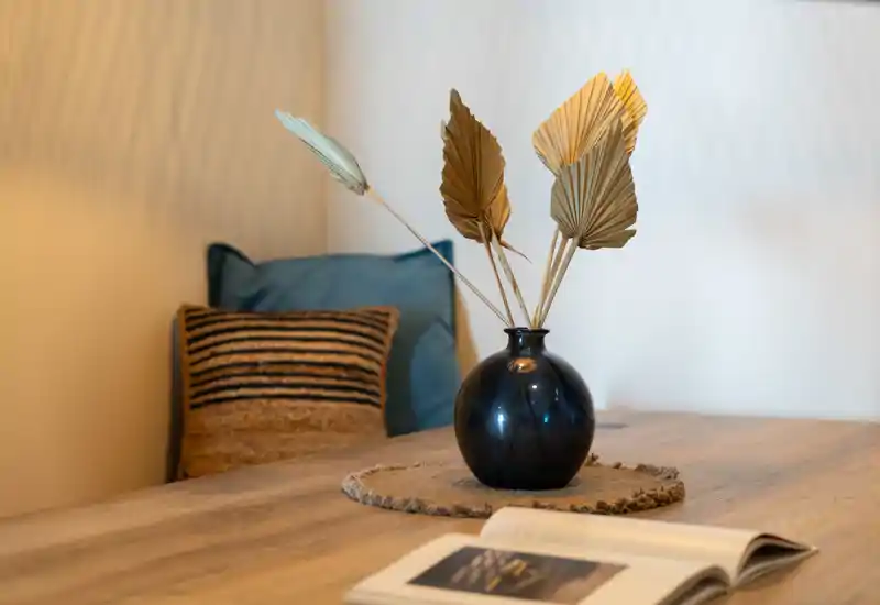 Primer plano de una mesa de salón decorada con un jarrón, hojas secas y cojines al fondo que crean un rincón acogedor.