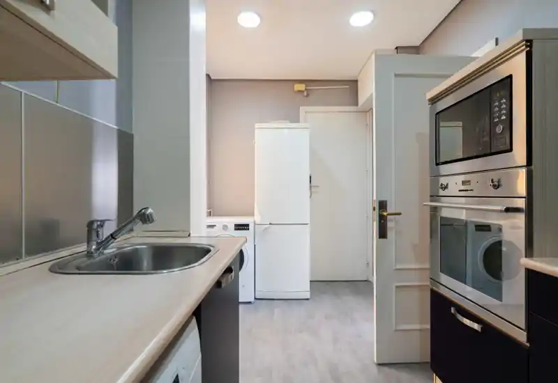 Cocina compacta y luminosa con electrodomésticos integrados: horno, microondas, frigorífico y fregadero de acero inoxidable. Líneas limpias y tonos neutros que ofrecen un espacio práctico y fácil de mantener, ideal para pisos urbanos.