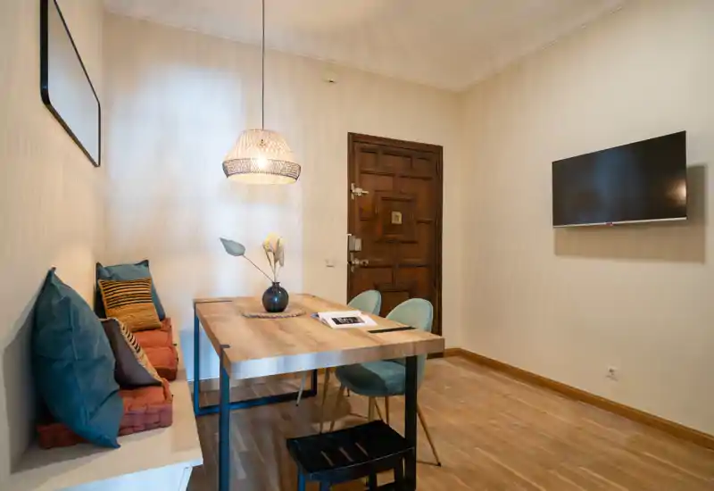 Agradable rincón comedor con mesa de madera, banco con cojines y dos sillas, iluminado por una lámpara colgante de mimbre y con TV en la pared — perfecto para comidas acogedoras o trabajar desde casa.