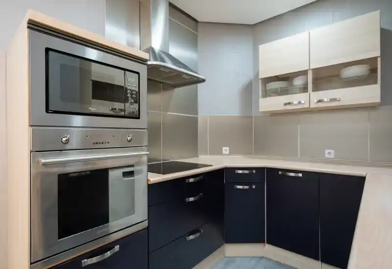 Cocina en esquina moderna con horno y microondas empotrados de acero inoxidable, muebles bajos oscuros y encimeras de madera clara; espacio limpio, bien iluminado y funcional, ideal para anuncios de alquiler.