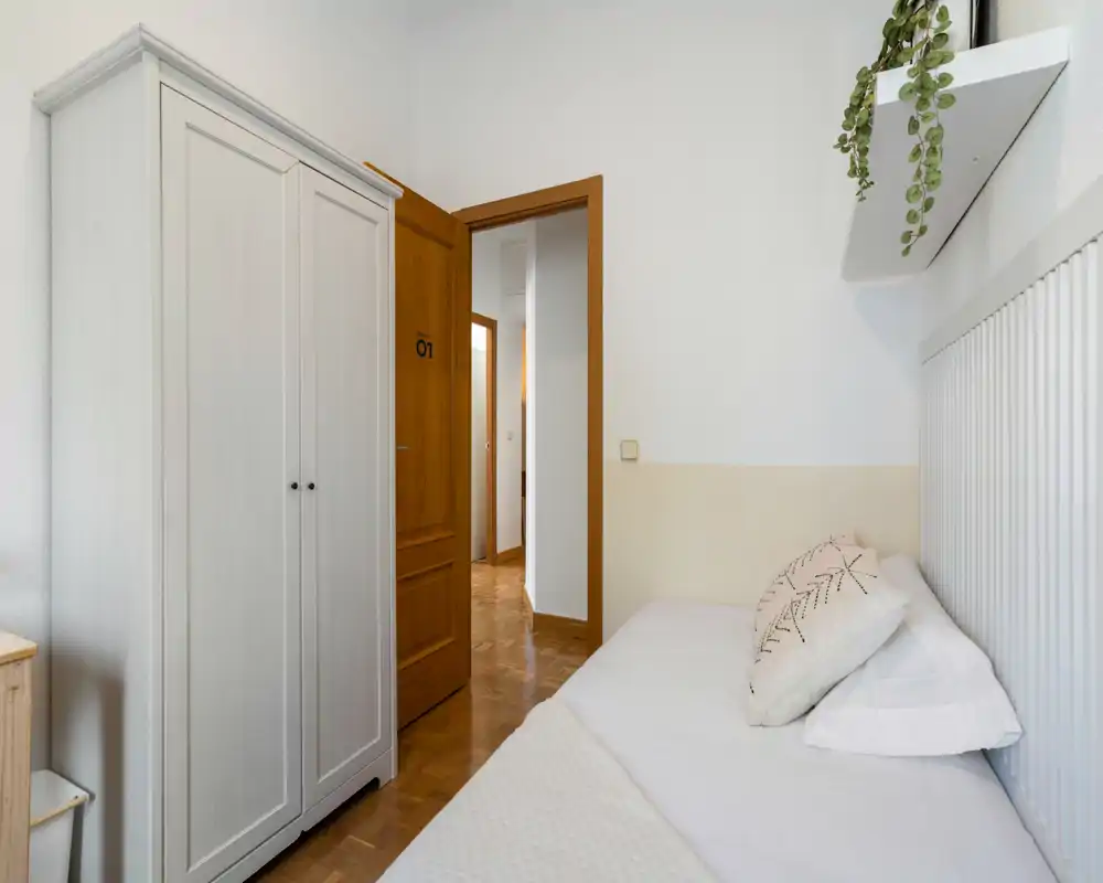 Dormitorio individual luminoso y ordenado con cama visible y textiles suaves; paredes blancas y puerta de madera que aportan una sensación minimal y tranquila.