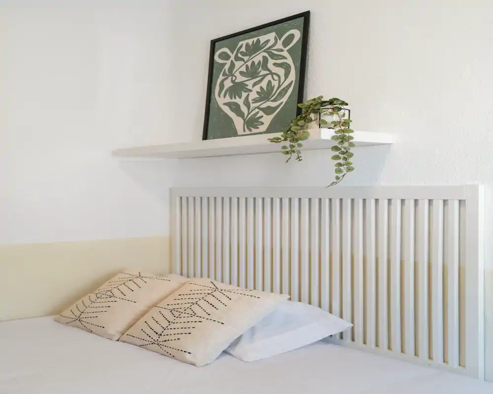 Rincón de dormitorio luminoso y minimalista con cabecero de listones, cojines decorativos, una repisa con cuadro y planta colgante: un área de descanso serena y bien decorada.