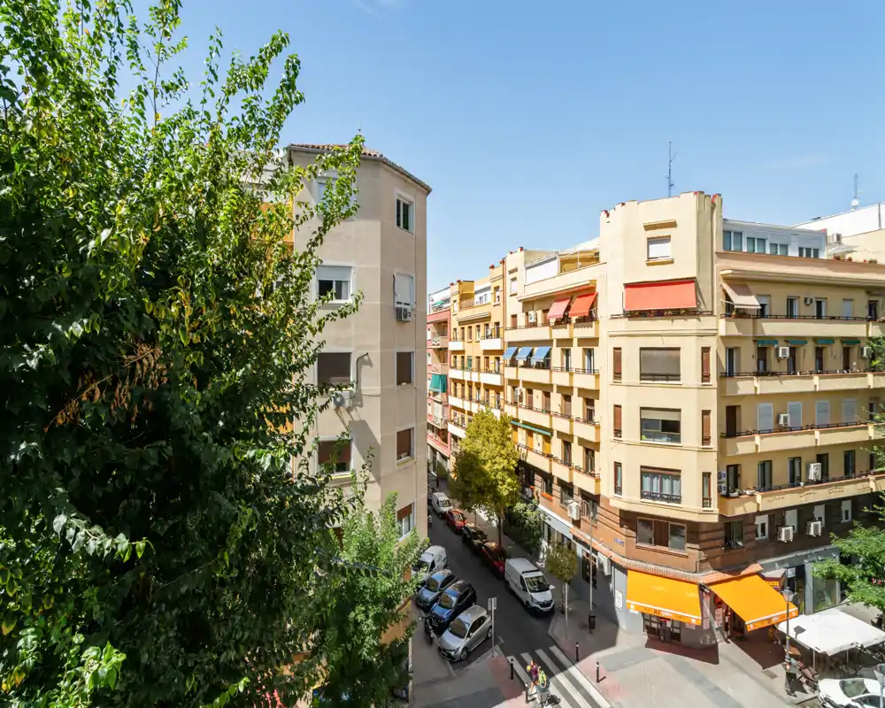 Vista soleada desde el balcón de una calle urbana con árboles, coches aparcados y edificios bajos — ideal para mostrar el barrio y los aspectos exteriores del anuncio.