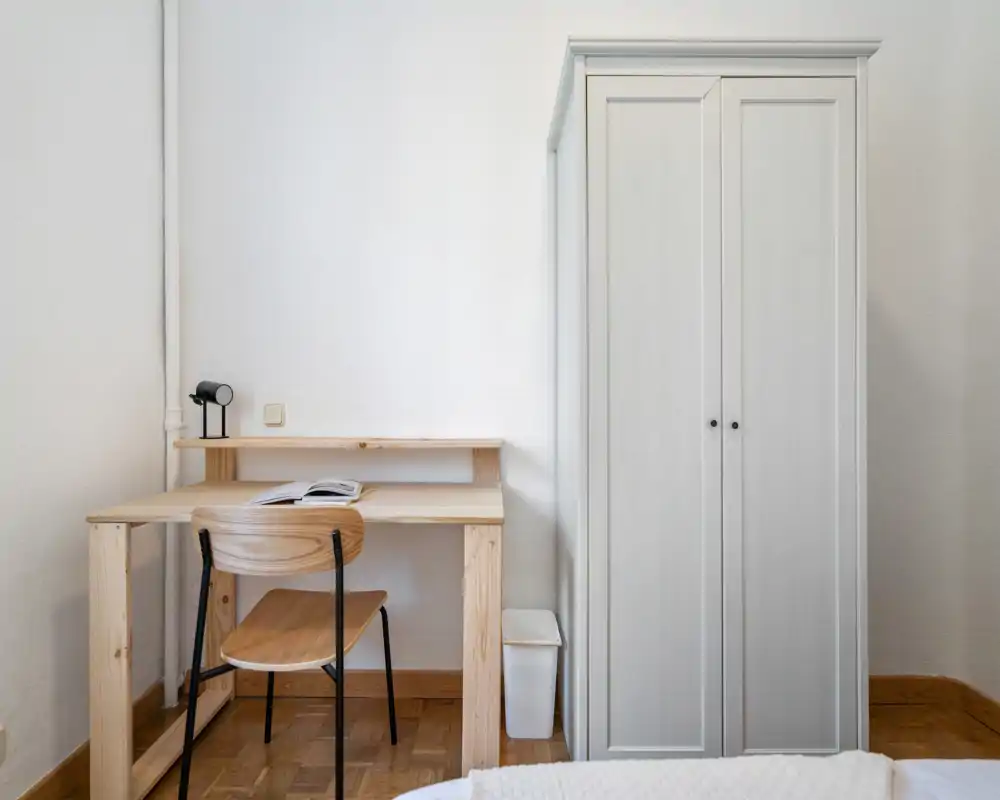 Rincón de dormitorio minimalista con un pequeño escritorio de madera, silla y un armario alto blanco; se ve un borde de la cama en primer plano. Espacio luminoso y despejado, ideal para trabajar y descansar.