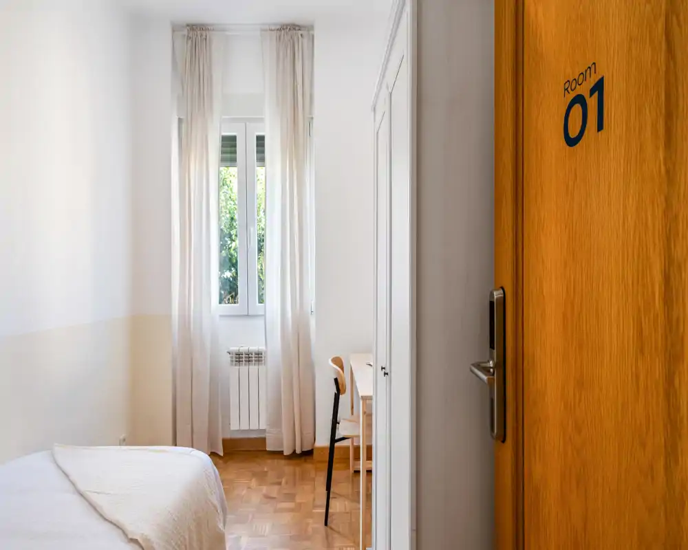 Habitación individual luminosa y minimalista con cama bien hecha, ventana alta con cortinas translúcidas, suelo de madera y un pequeño escritorio — ambiente tranquilo y aireado ideal para estancias cortas.