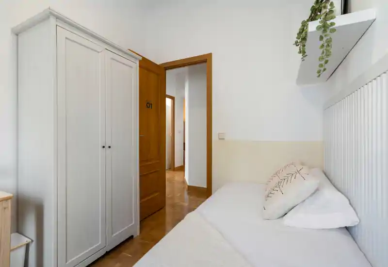 Bright, neatly styled single bedroom with a visible daybed and soft textiles; white walls and wooden door create a calm, minimalist feel.