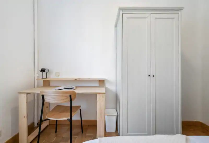 Rincón de dormitorio minimalista con un pequeño escritorio de madera, silla y un armario alto blanco; se ve un borde de la cama en primer plano. Espacio luminoso y despejado, ideal para trabajar y descansar.