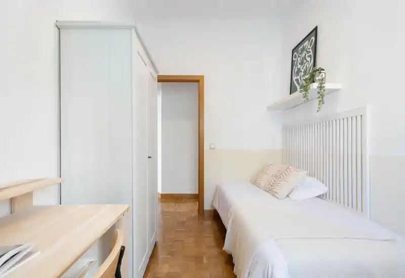 Bright, minimal single bedroom with a tidy bed, wooden parquet floor, a wardrobe and a small desk — calm, airy and well-styled for rental listings.
